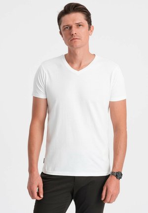 Biały bawełniany T-shirt z dekoltem w serek, krótki rękaw, regularny krój, gładka tekstura. Noszony z ciemnymi spodniami; posiada małą metkę marki na dolnym brzegu.