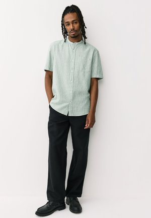 Homme avec des dreadlocks portant une chemise à manches courtes vert clair boutonnée, pantalon noir et chaussures noires, debout avec une main dans la poche.