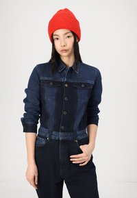 Denim jakna v kontrastnih odtenkih temno in svetlo modre, z gumbnim zapiranjem spredaj, žepoma na prsih in strukturirano silhueto. Rdeča pletena kapa.
