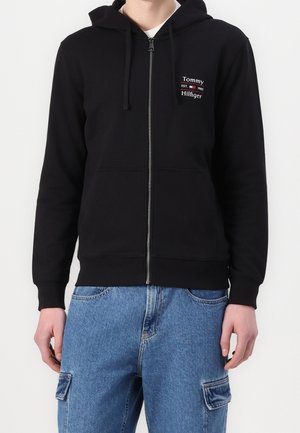 Person trägt schwarzen Hoodie mit Reißverschluss und "Tommy Hilfiger"-Logo auf der Brust und blaue Cargo-Jeans, steht vor weißem Hintergrund.