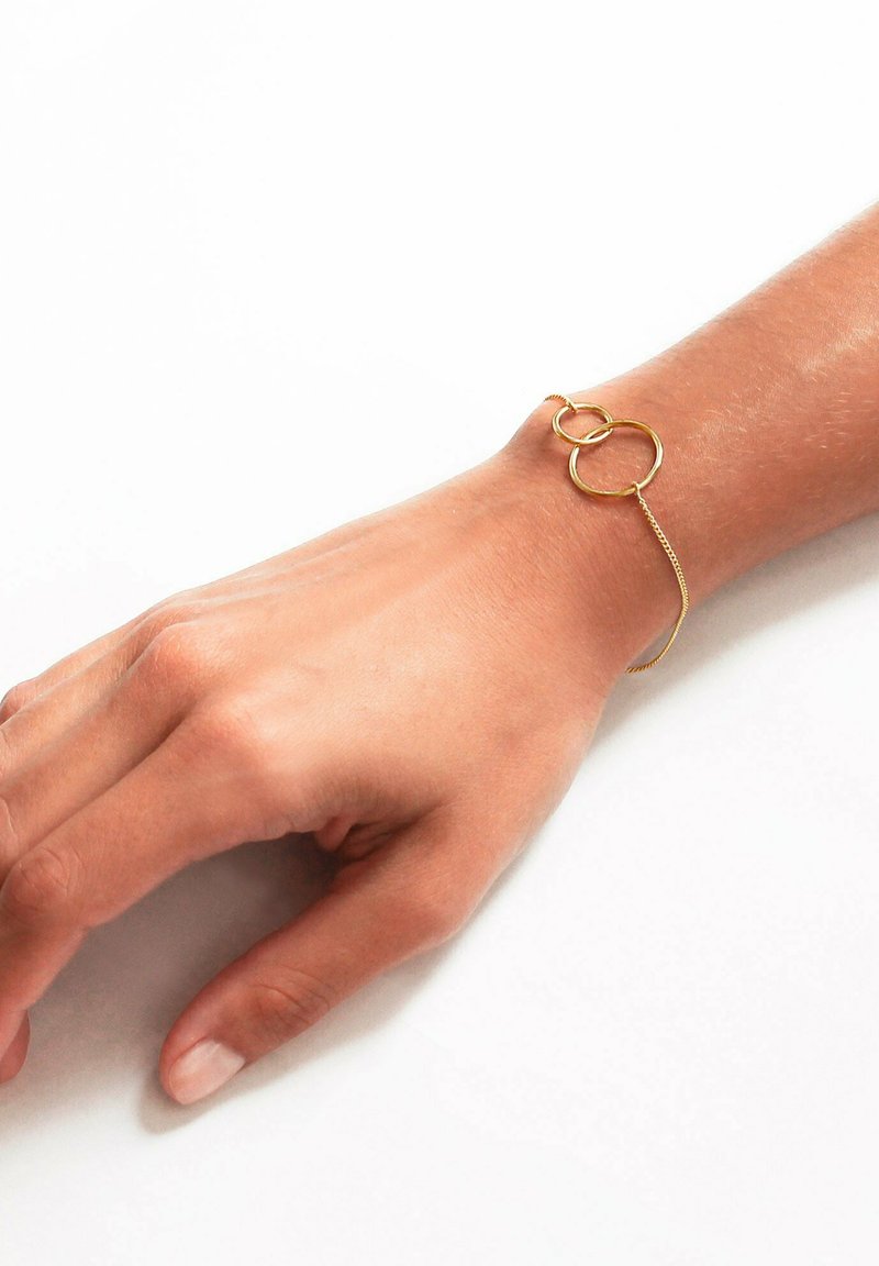 Bracciale d'oro con due anelli circolari interconnessi, accompagnato da una delicate catena. Il design è minimalista ed elegante contro il tono della pelle.