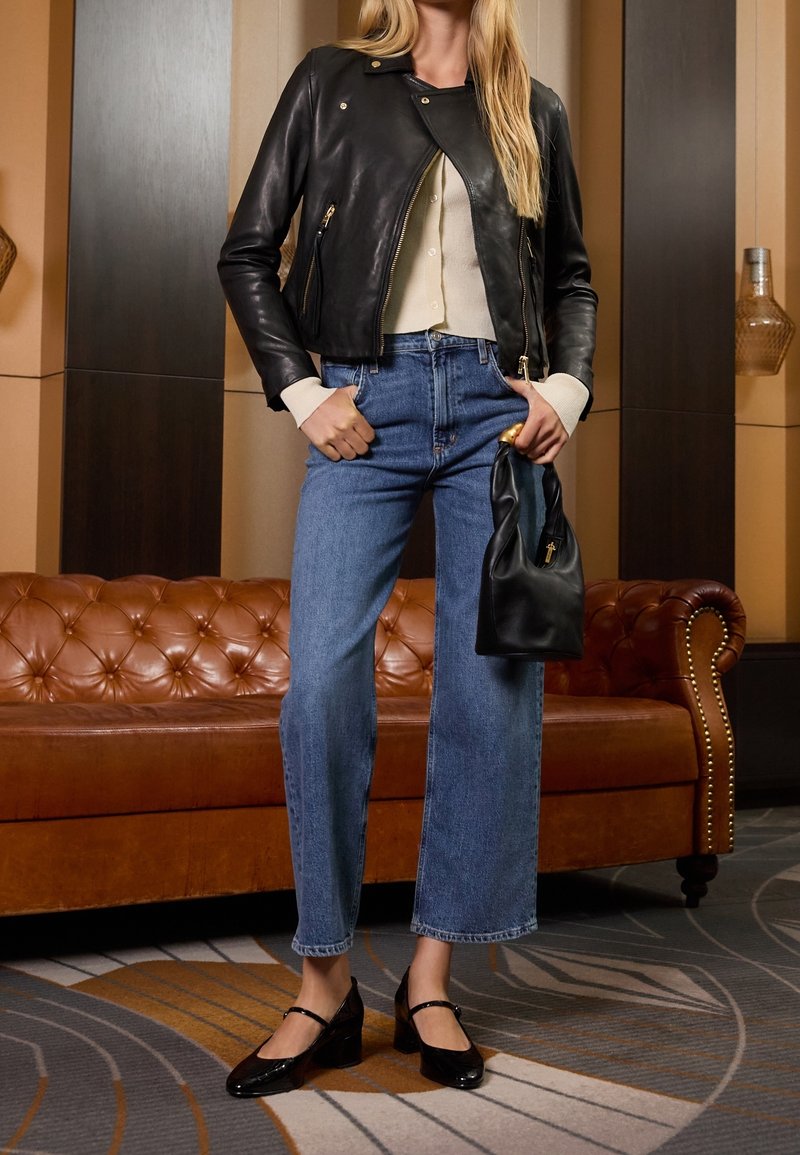 Femme portant une veste en cuir noire, un cardigan crème, un jean bleu taille haute, des talons noirs, tenant un petit sac à main noir à l'intérieur.