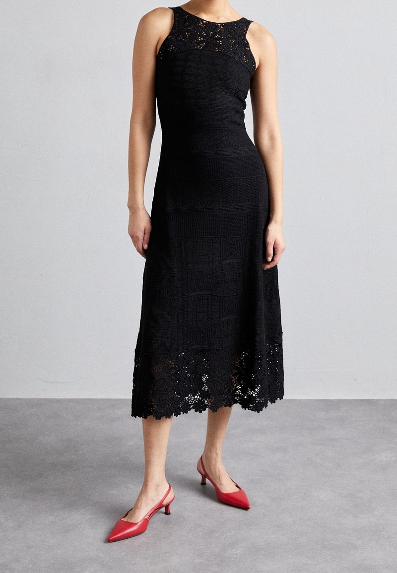 Femme portant une robe midi noire en dentelle sans manches avec une taille cintrée et des talons slingback rouges à bout pointu, debout sur un sol gris.