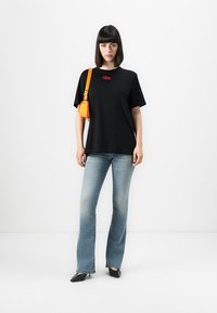 Woman with straight dark hair wearing black oversized t-shirt, faded flare jeans, black heels, and carrying a small orange shoulder bag.