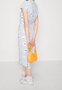 Ljusblå klänning med vitt blommönster, korta ärmar, åtsittande midja och knälängd. Håller en liten texturerad gul handväska. Vita sneakers.