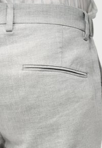 Pantaloni din material gri deschis, cu buzunar dublu la spate și o buclă de curea vizibilă, purtați cu o cămașă în dungi băgată în pantaloni.