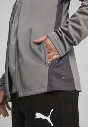 Graue Sportjacke mit strukturierten Einsätzen, Reißverschluss und Taschen. "DRYCELL"-Label auf der Unterseite, kombiniert mit schwarzen Hosen mit weißem Logo.