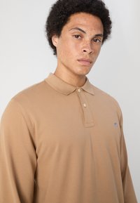 Polo de manga larga en beige, con un cuello clásico, tres botones y un logo bordado en el lado izquierdo del pecho. Textura de tela suave.