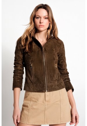 Junge Frau mit langen hellbraunen Haaren, die eine braune Wildlederjacke mit Reißverschluss und einen beigen Rock trägt, steht vor weißem Hintergrund.