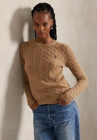 Femme aux cheveux tressés portant un pull en maille torsadée couleur camel et un jean bleu, debout les mains dans les poches devant un mur uni.