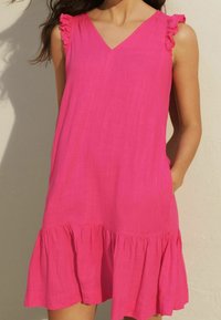 Vestido sin mangas rosa con escote en V, hombros con volantes y dobladillo de volantes en capas. Fabricado con una tela ligera y con bolsillos laterales.