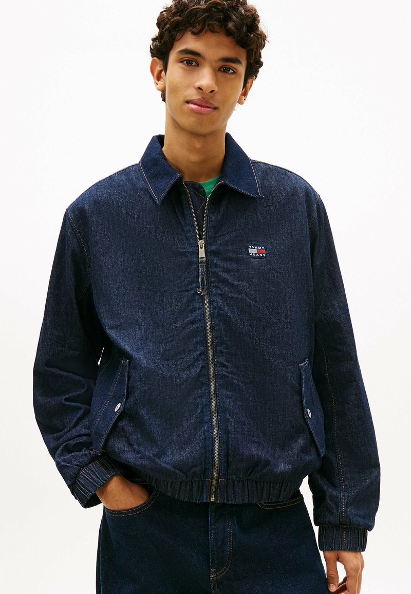 Jeune homme portant une veste en denim bleu foncé à fermeture éclair avec des poches à boutons et un logo Tommy Jeans sur la poitrine, les mains dans les poches.