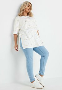 Top blanco y holgado con rayas doradas y grises, con aberturas laterales, combinado con jeans ajustados azul claro y zapatillas blancas.