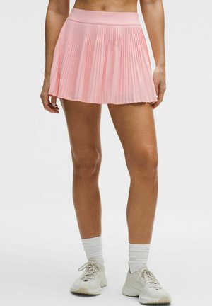 Mujer con una falda plisada rosa, calcetines blancos hasta el tobillo y zapatillas deportivas blancas, de pie frente a un fondo liso.