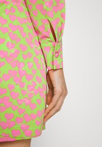 Vestido curto em tecido verde brilhante com um padrão de corações rosa, detalhe de punhos abotoados e uma fenda lateral.