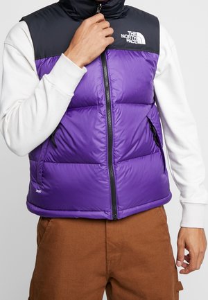 Man die een paars-zwarte mouwloze donsvest met wit logo, wit shirt met lange mouwen en een bruine broek draagt, en de rits van het vest verstelt.