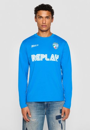 Man wearing a bright blue long-sleeve shirt with "REPLAY" logo and crest, paired with light blue distressed jeans against a plain background.