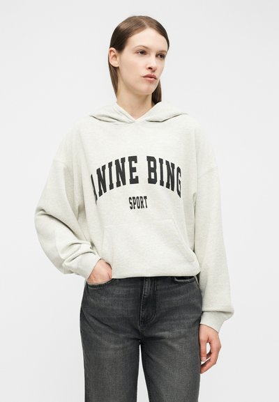 Woman wearing light gray hoodie with "ANINE BING SPORT" text, paired with dark gray jeans, standing against a plain white background.