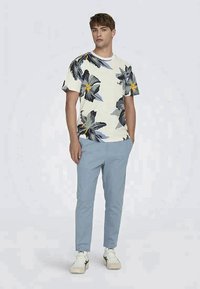 Lightweight, cream T-shirt with large floral print in gray, yellow, and blue, paired with light blue trousers and white sneakers.