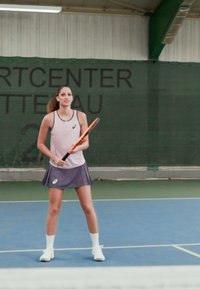 Tenisašica u svijetloružičastom topu bez rukava i tamnoj suknji, drži narančasti teniski reket, na plavom terenu s pozadinom zelene mreže.