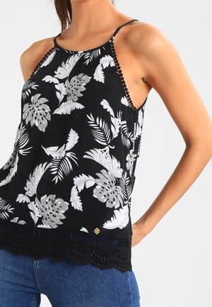 Femme portant un haut noir sans manches avec un motif de feuilles tropicales blanches et un ourlet en dentelle crochetée, associé à un jean bleu, sur fond neutre.