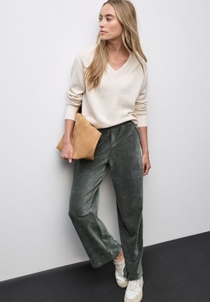 Pull beige à col en V, pantalon large en velours vert, tenant une pochette en suède beige, et portant des baskets blanches sur un fond uni.