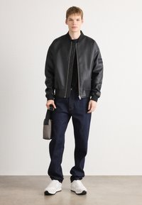 Chaqueta bomber de cuero sintético negro con puños acanalados, combinada con jeans de mezclilla oscura y zapatillas blancas. Modelo sosteniendo un pequeño bolso gris.