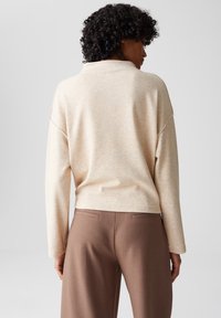 Vrouw met krullend haar, gekleed in een beige longsleeve en bruine broeken, staat met haar rug naar de camera tegen een effen achtergrond.