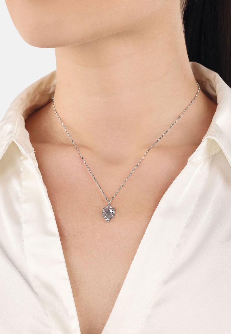 Silverhalsband med en delikat kedja som har en glittrande, facetterad sten i en kvadratisk infattning, uppvisad mot en vit skjorta.