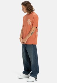Hombre con pelo rizado de longitud media que lleva una camiseta gráfica naranja, jeans azules holgados, zapatillas blancas y un tatuaje de tiburón en su antebrazo.