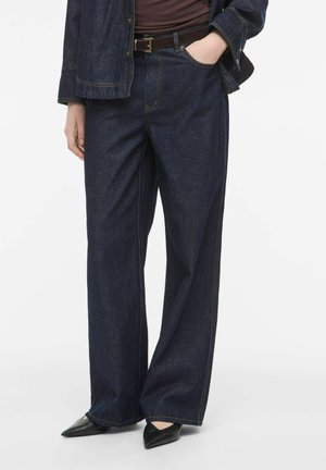 Persoon die donkerblauwe wijde jeans draagt, zwarte puntige schoenen, een bruine riem en een bijpassend donkerblauw spijkerjasje, één hand in de zak.