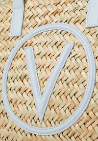 Nertinė šiaudų pirkinių krepšys su dideliu šviesiai pilku odiniu "V" logotipu ir kontrastingais smėlio bei geltonais austais tekstūromis.