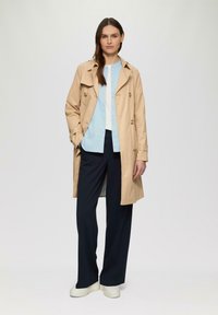 Beige trenchcoat met dubbelzijdige knopen, lichtblauw shirt, marineblauwe wijde broek en witte platform sneakers op een neutrale achtergrond.
