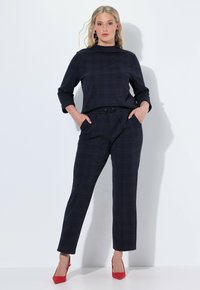 Blonďatá žena stojí s rukama v kapsách, oblečená v námořnickém kostkovaném dvoudílném obleku a červených vysokých podpatcích, pózující na bílém pozadí.