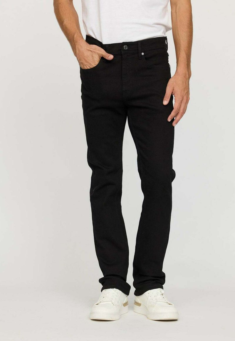 Homme portant un jean noir slim et des baskets blanches, avec la main gauche dans la poche et la main droite détendue sur le côté.