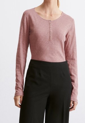 Nainen pukeutunut pölyisen vaaleanpunaiseen pitkähihainen henley-paitaan, joka on laitettu korkeavyötäröisten mustien housujen sisään, seisoo yksiväristä valkoista taustaa vasten.