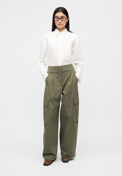 Λευκό πουκάμισο με γιακά και μακριά μανίκια, συνδυασμένο με φαρδιά χακί cargo παντελόνια με πλαϊνές τσέπες. Το μοντέλο φορά καφέ παπούτσια.