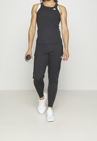 Débardeur de sport noir avec des accents blancs, associé à un pantalon de jogging noir fuselé avec des panneaux texturés, et des baskets blanches. Bouteille d'eau à la main.