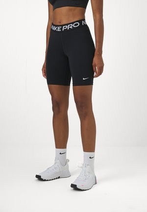 Crne Nike Pro kratke hlače s širokim brendiranim pojasom, uskim dizajnom i glatkom teksturom, uparenim sa bijelim sportskim cipelama i čarapama.