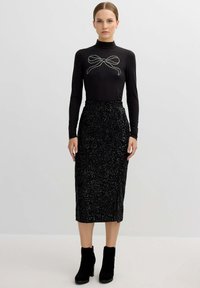 Top negro de manga larga con un detalle de lazo de strass, combinado con una falda de lentejuelas negras hasta la rodilla. Botines negros de tacón completan el look.