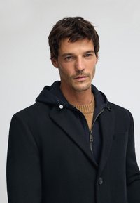 Svart kappa i ullblandning med huva, figursydd design och subtil textur, lager-på-lager över en brun stickad tröja med dragkedja.