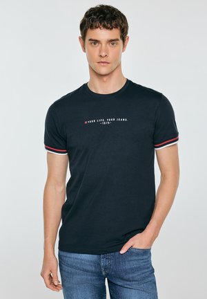 Młody mężczyzna w czarnej koszulce z czerwono-białymi paskowanymi rękawami i niebieskich dżinsach, stoi z jedną ręką w kieszeni na jednolitym tle.