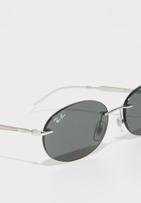 Ray-Ban ovalios juodos lęšiai saulės akiniai su plonu sidabro metaliniu rėmeliu ir skaidriais nosies pagalvėlėmis ant balto fono.