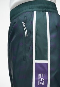 Nylon-Shorts in Dunkelgrün mit weißen Seitenstreifen, einem elastischen Bund, einer zippten Tasche und lila EA7-Logo-Detail.