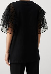 Camiseta negra con mangas de lunares transparentes y volantes. La tela es de algodón suave, mientras que las mangas presentan un diseño aireado y en capas.