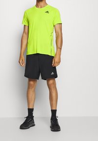 Neon gul idrottströja med svarta detaljer, rund halsringning; matchad med svarta shorts och svarta sportskor.