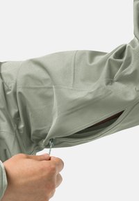 Lichtgroene jassenstof met een gladde textuur; toont een zijzak met rits die door een hand wordt geopend. Stikdetails zichtbaar.