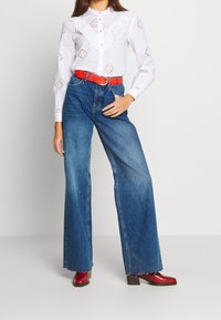 Chemise blanche boutonnée avec détails en œillets, jean bleu à jambes larges, ceinture rouge en simili cuir et bottines rouges. Tissus lisses et texturés.