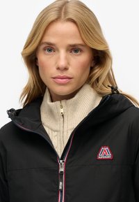 Sort hættejakke med en rød og blå detaljeret lynlås, prydet med et logo-patch. Båret over en lys cremefarvet ribbestrikket sweater med høj krave.