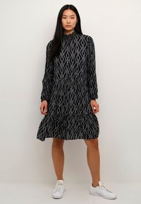 Zwarte jurk met wit golfpatroon, hoge halslijn, lange mouwen en gelaagde uitvoering, gecombineerd met witte sneakers. Gladde textuur.